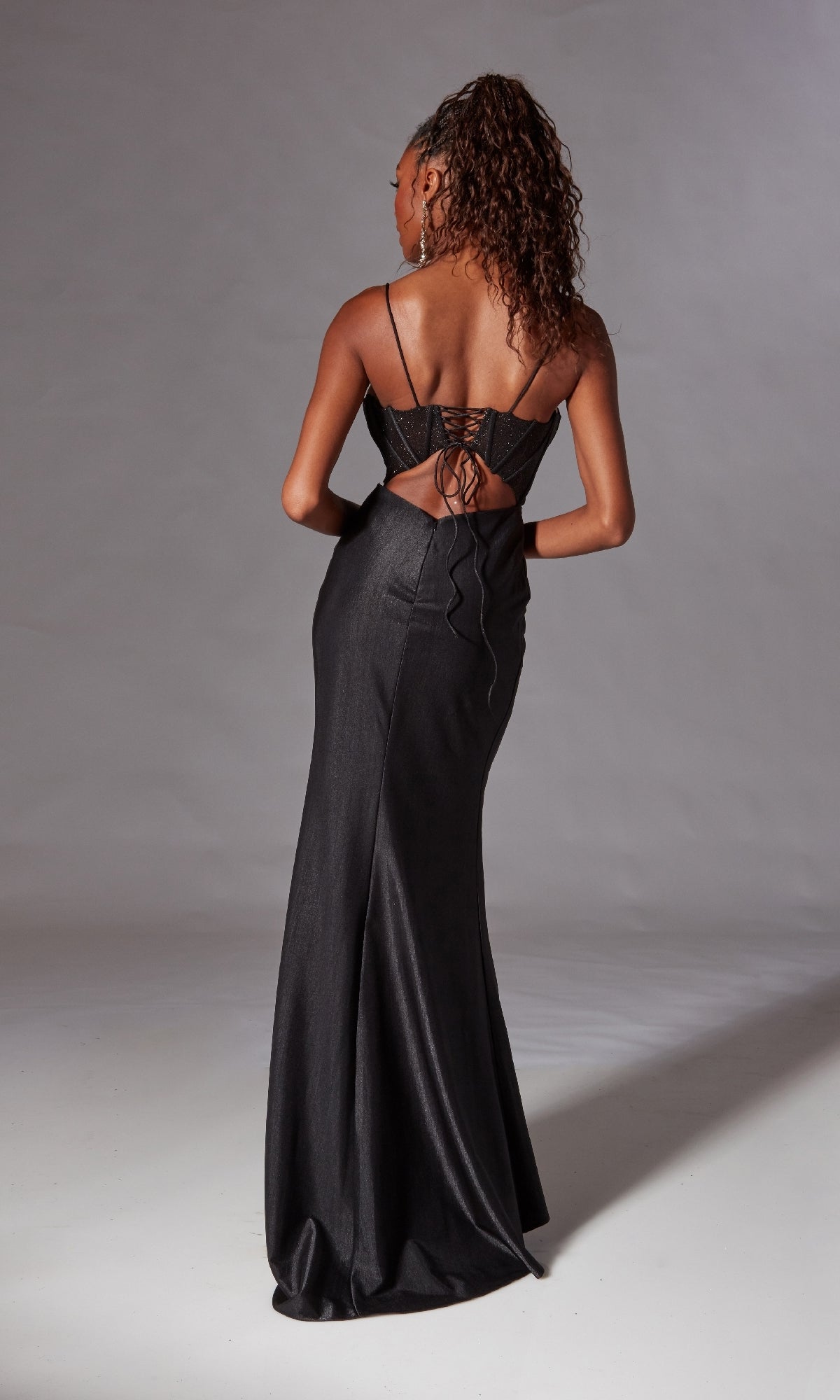 A woman faces away, wearing the Aerie Couture 269 corset-bodice long prom dress—black, glittery, with thin straps and an open lace-up back. Her curly hair is styled in a high ponytail.